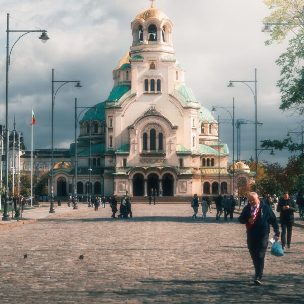 People walk along a cobblestone path leading to the large, ornate Alexander Nevsky Cathedral in Sofia, Bulgaria, under a cloudy sky. The cathedral features golden domes and green rooftops.